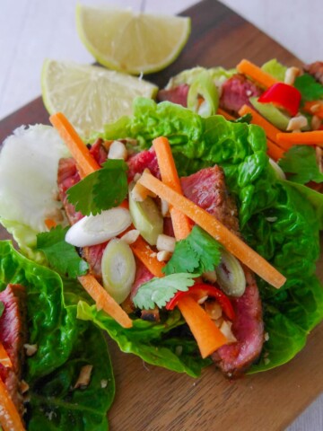 A wooden board topped with 3 lettuce leaves, laden with sliced lemongrass steak strips, crunchy carrot strips, spring onion, crushed peanuts and coriander leaf with wedges of fresh lime.