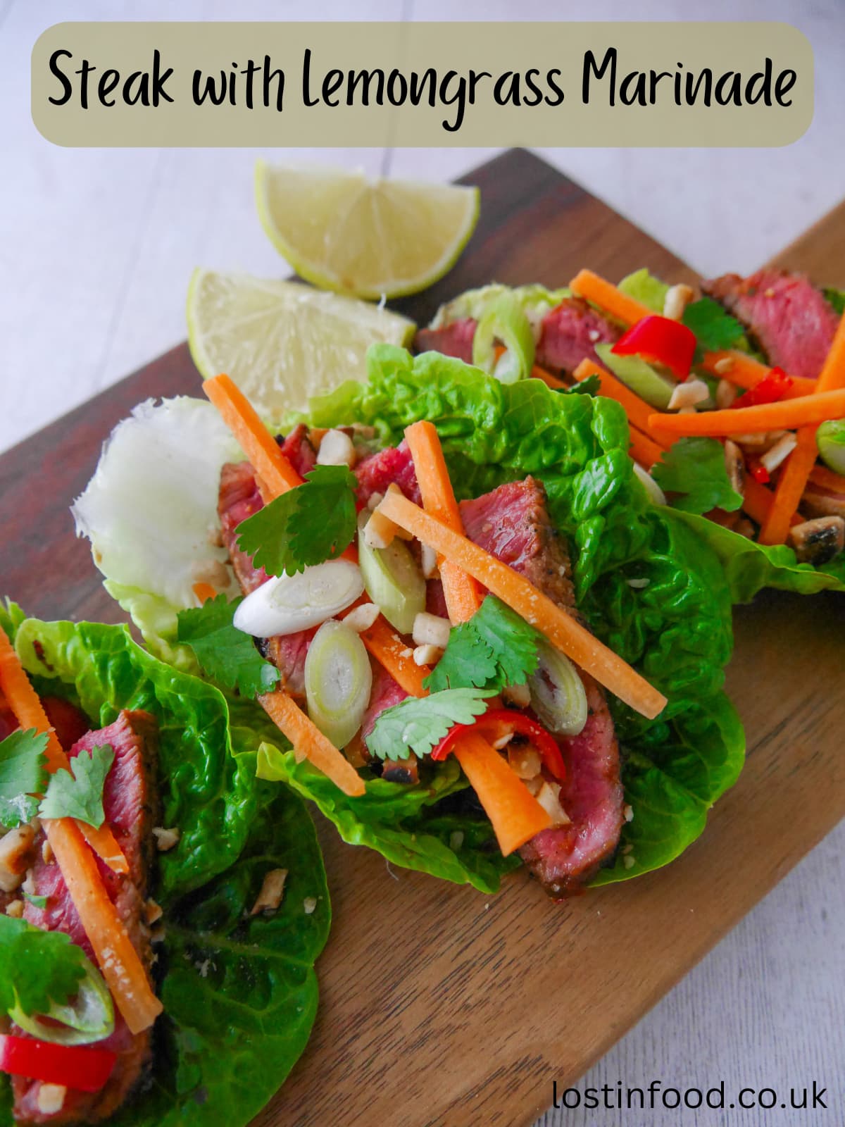 Pinnable image with recipe title and a wooden board topped with 3 lettuce leaves, laden with sliced lemongrass steak strips, crunchy carrot strips, spring onion, crushed peanuts and coriander leaf with wedges of fresh lime.