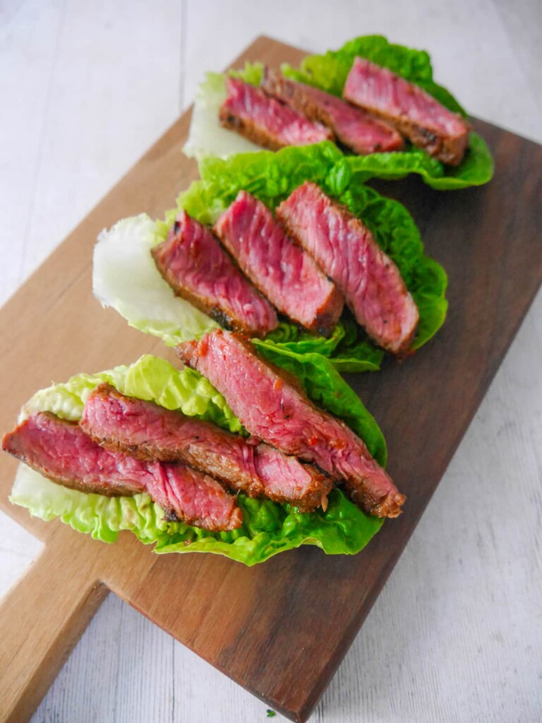 A wooden board with lettuce leaves, topped with thin slices of cooked sirloin.