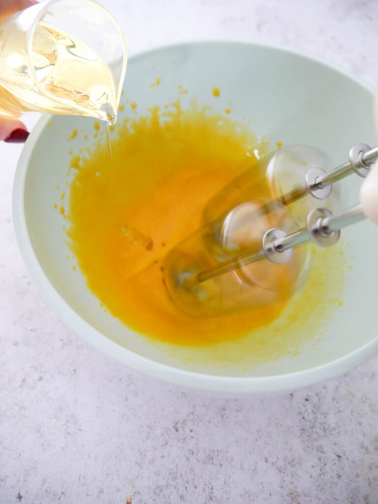 Sunflower oil being slowly poured into a bowl of egg yolks and mustard being whisked with an electric hand mixer.