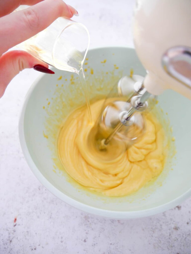 Sunflower oil being slowly poured into a bowl of egg yolks and mustard being whisked with an electric hand mixer.