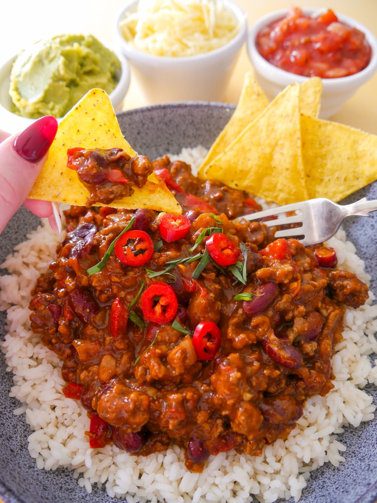 A bowl of steamed rice, topped with minced beef chilli, with bowls of guacamole, shredded cheese and tomato salsa set alongside.