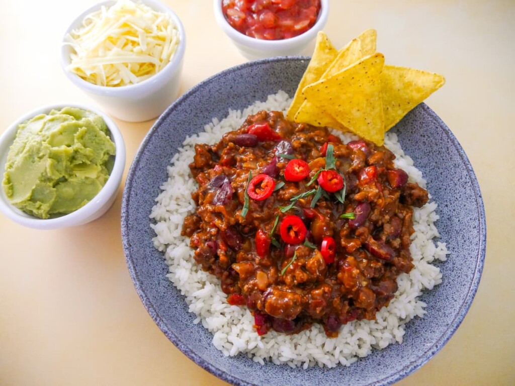 A bowl of steamed rice, topped with minced beef chilli, with bowls of guacamole, shredded cheese and tomato salsa set alongside.