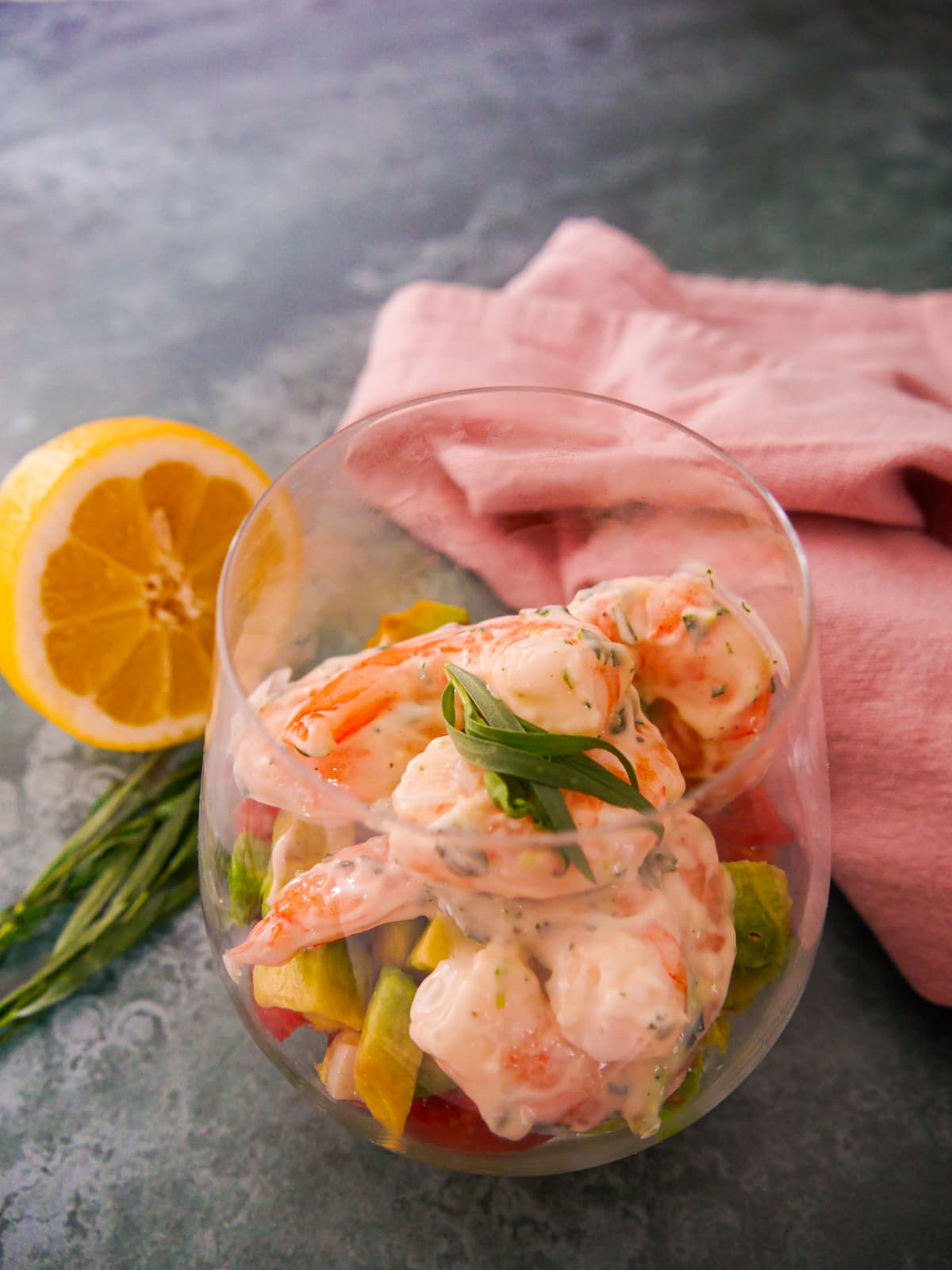 A large glass filled with a base of shredded lettuce, chopped tomato and avocado, topped with king prawns in a tarragon mayonnaise, garnished with fresh tarragon.
