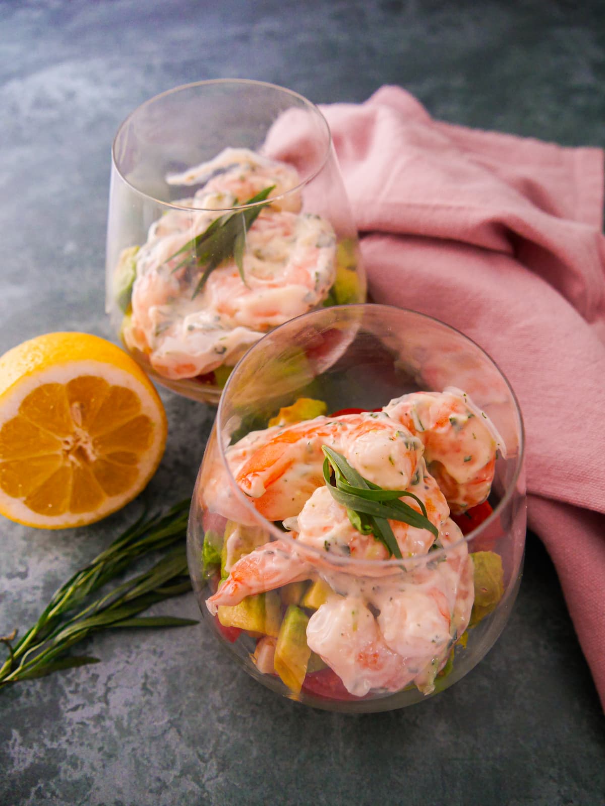 Two large glasses filled with a base of shredded lettuce, chopped tomato and avocado, topped with king prawns in a tarragon mayonnaise, garnished with fresh tarragon.