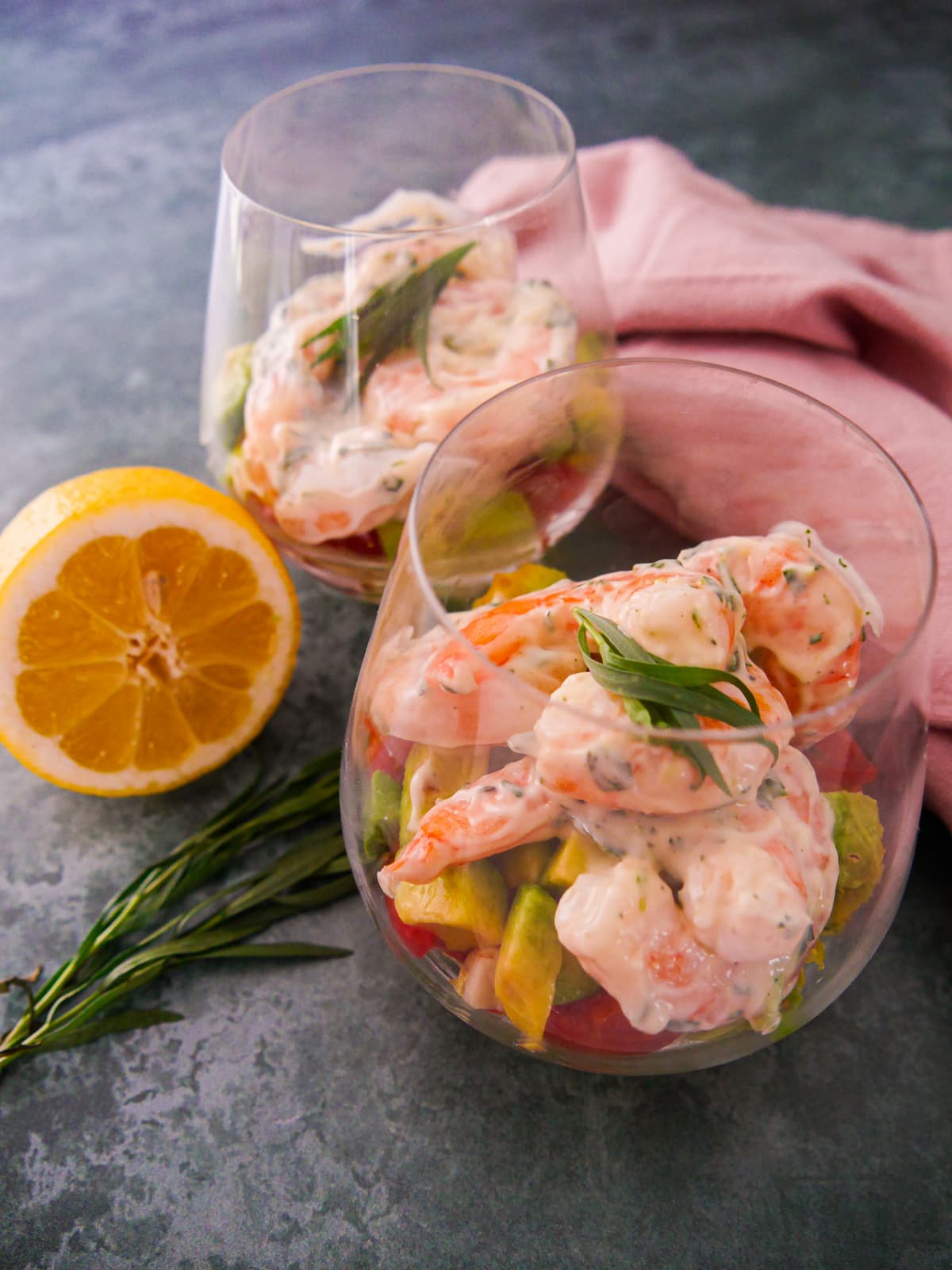 Two large glasses filled with a base of shredded lettuce, chopped tomato and avocado, topped with king prawns in a tarragon mayonnaise, garnished with fresh tarragon.