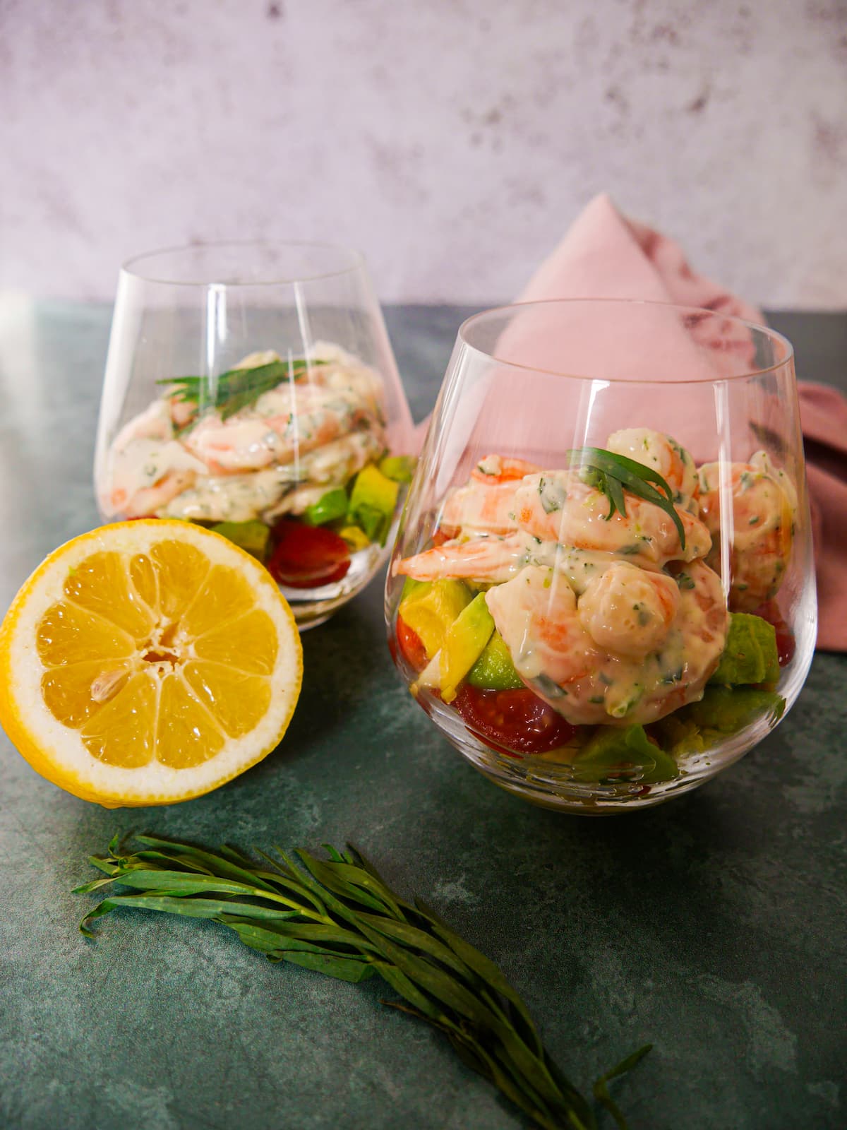 Two large glasses filled with a base of shredded lettuce, chopped tomato and avocado, topped with king prawns in a tarragon mayonnaise, garnished with fresh tarragon.