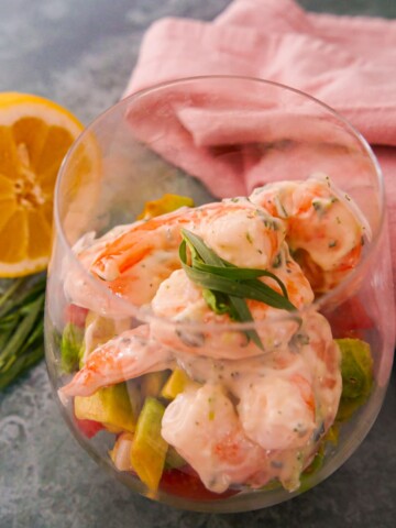 A large glass filled with a base of shredded lettuce, chopped tomato and avocado, topped with king prawns in a tarragon mayonnaise, garnished with fresh tarragon.