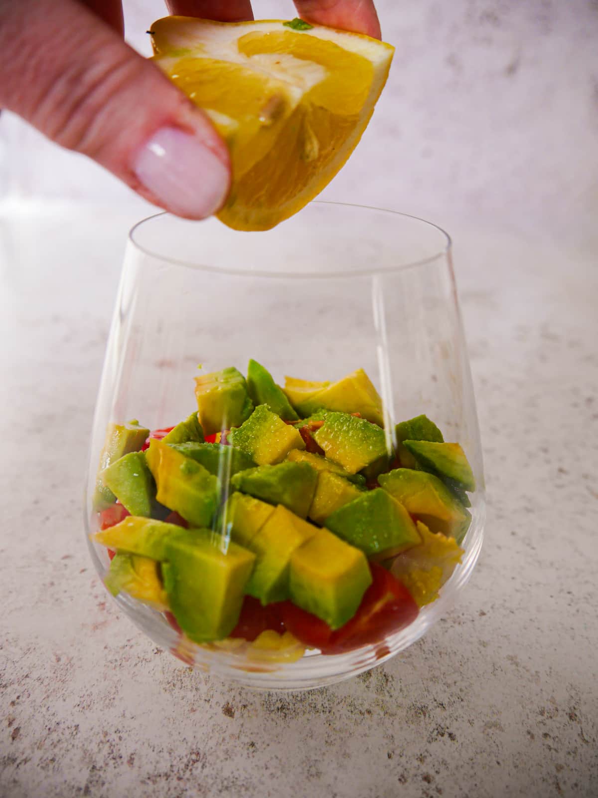 A large glass filled with a base of shredded lettuce, chopped tomato and avocado, with fresh lemon juice being squeezed over.