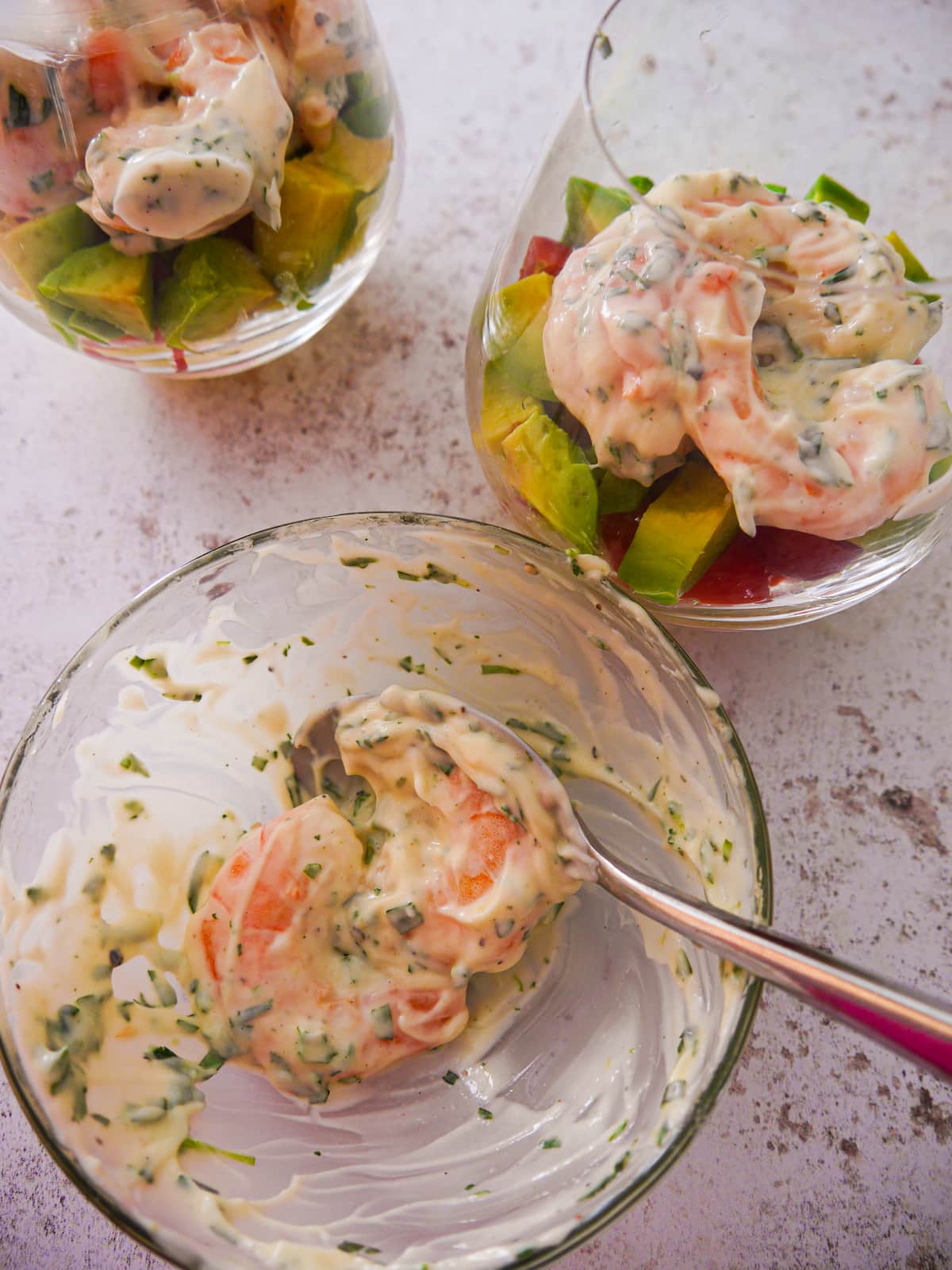 A bowl of cooked king prawns being covered with tarragon mayonnaise, with two glasses of prawn cocktail set alongside.