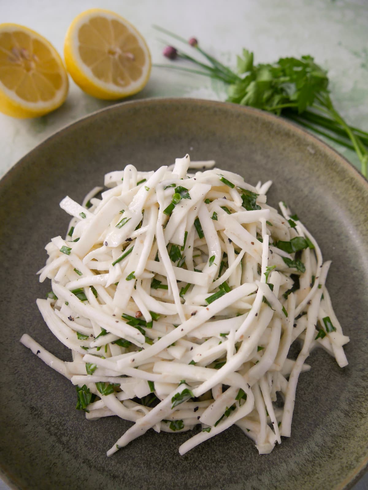 A green bowl of celeriac remoulade salad, with lemon and fresh parsley and chives set alongside.