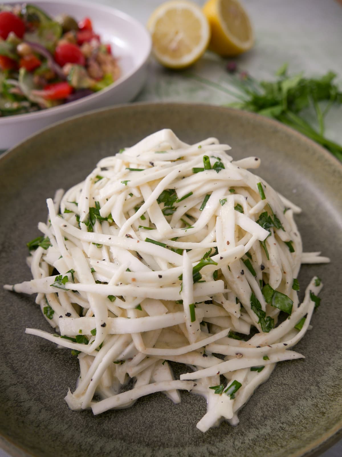 A green bowl of celeriac remoulade salad, with lemon and fresh parsley and chives set alongside.