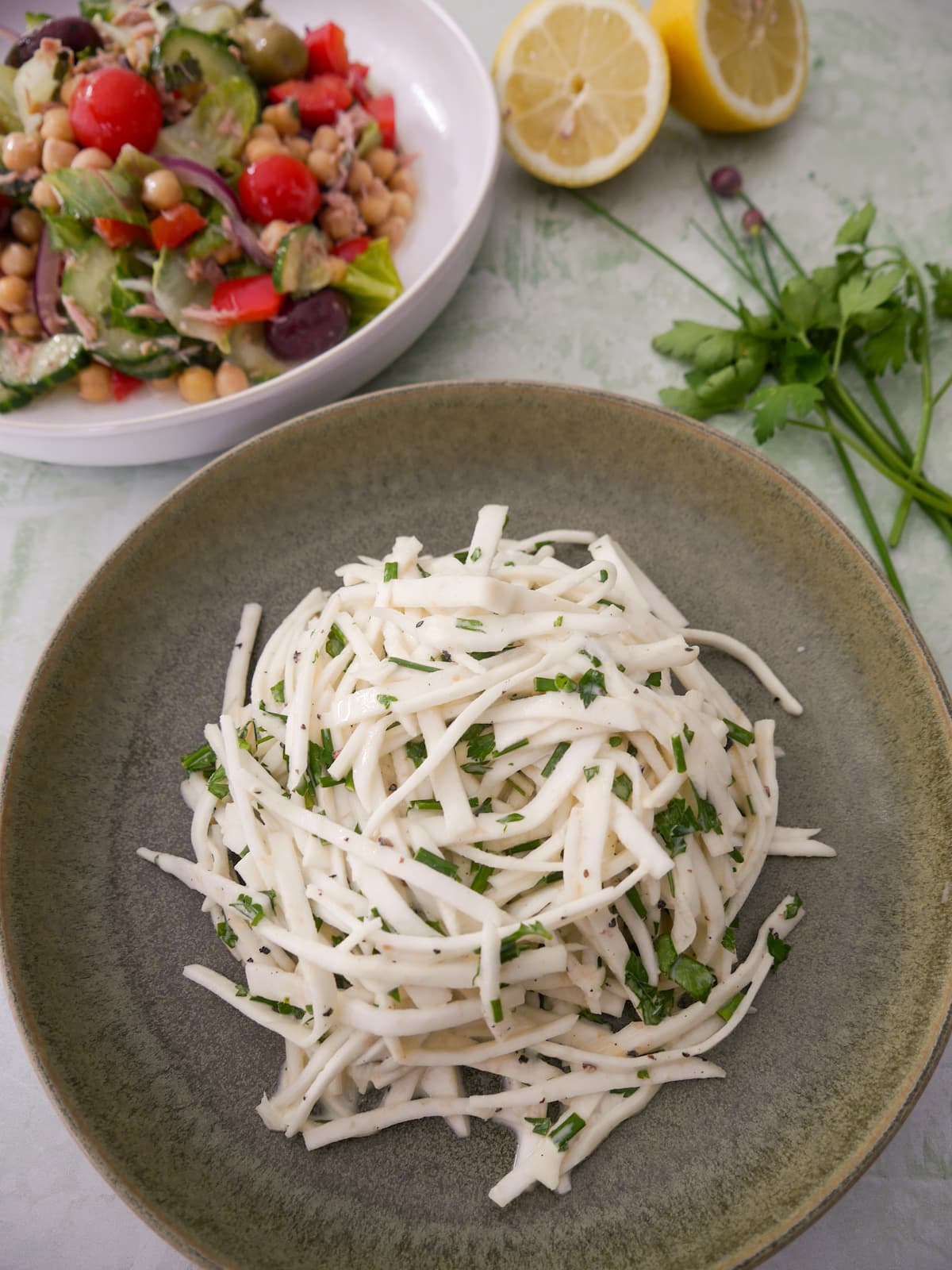 A green bowl of celeriac remoulade salad, with lemon and fresh parsley and chives set alongside, and a bowl of mixed vegetable and chickpea salad set alongside.