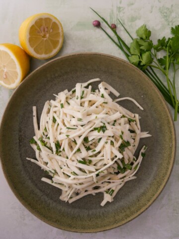 A green bowl of celeriac remoulade salad, with lemon and fresh parsley and chives set alongside.