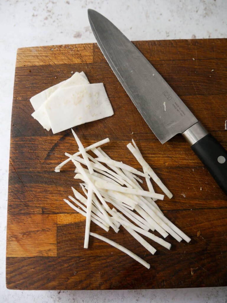 A chopping board with thin slices of celeriac being cut into thin strips.