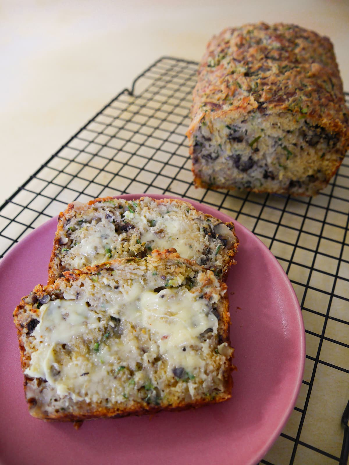 Courgette and cheese bread with walnuts on a wire cooling rack with a plate containing two butter slices of the bread.
