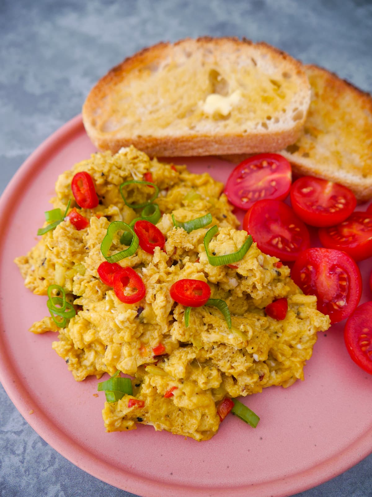 A pink plate topped with curried scrambled eggs with a garnish of thinly sliced red chilli and spring onion, served with 2 sliced of toast and some cherry tomato halves.