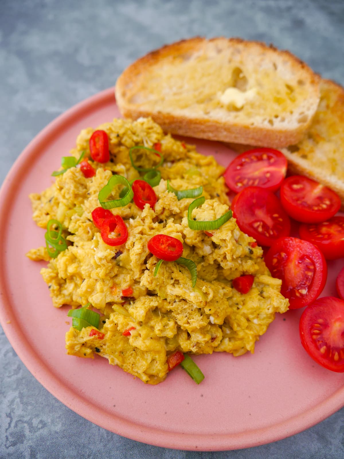 A pink plate topped with curried scrambled eggs with a garnish of thinly sliced red chilli and spring onion, served with 2 sliced of toast and some cherry tomato halves.