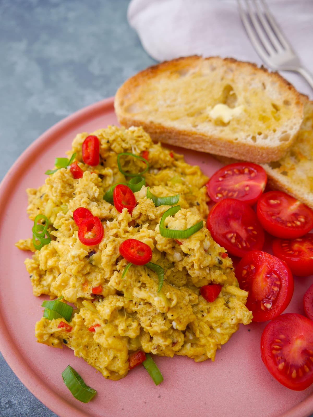 A pink plate topped with curried scrambled eggs with a garnish of thinly sliced red chilli and spring onion, served with 2 sliced of toast and some cherry tomato halves.
