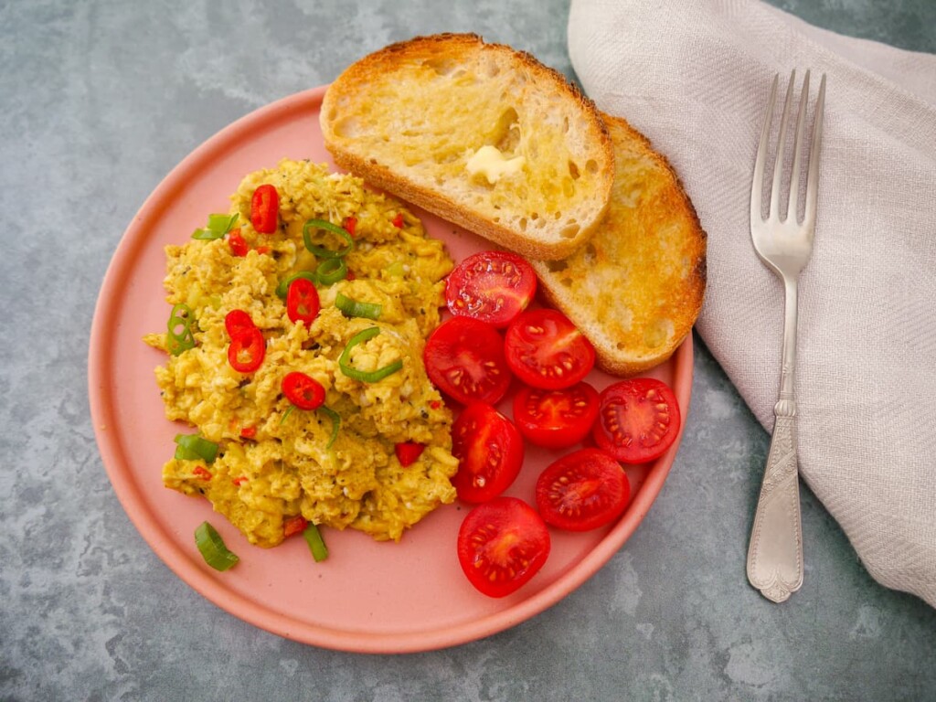A pink plate topped with curried scrambled eggs with a garnish of thinly sliced red chilli and spring onion, served with 2 sliced of toast and some cherry tomato halves.