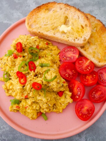 A pink plate topped with curried scrambled eggs with a garnish of thinly sliced red chilli and spring onion, served with 2 sliced of toast and some cherry tomato halves.
