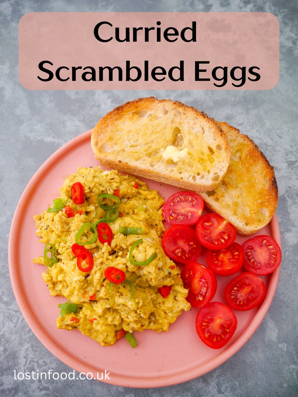 Pinnable image with recipe title and a pink plate topped with curried scrambled eggs with a garnish of thinly sliced red chilli and spring onion, served with 2 sliced of toast and some cherry tomato halves.