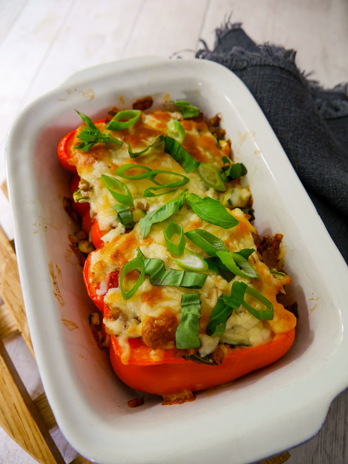 An oven proof dish with 2 halves of stuffed red pepper topped with melted cheese and a garnish of sliced spring onion and shredded basil leaves.