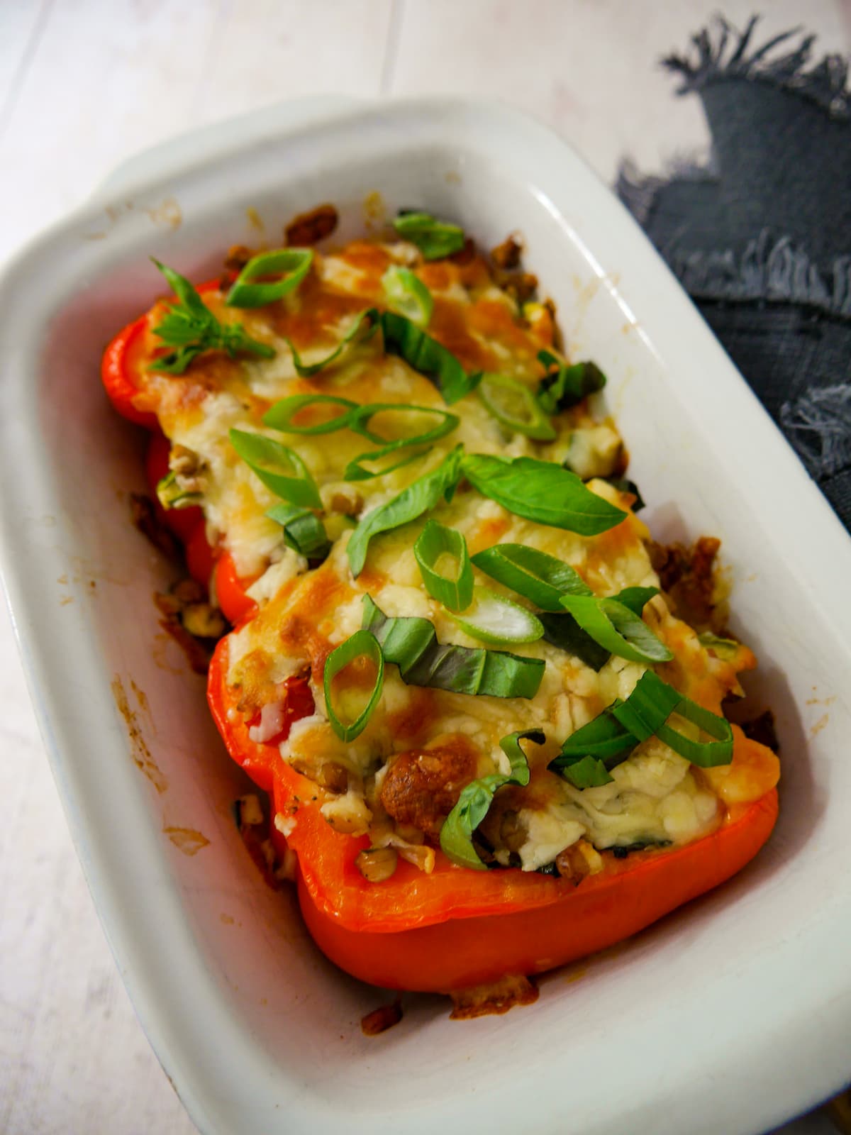 An oven proof dish with 2 halves of stuffed red pepper topped with melted cheese and a garnish of sliced spring onion and shredded basil leaves.