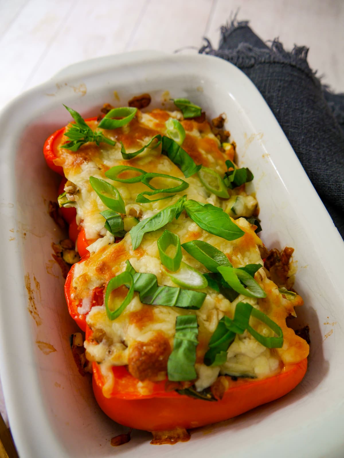 An oven proof dish with 2 halves of stuffed red pepper topped with melted cheese and a garnish of sliced spring onion and shredded basil leaves.
