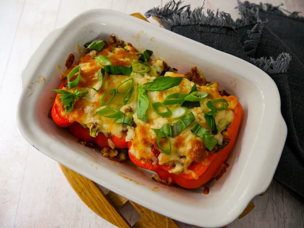 An oven proof dish with 2 halves of stuffed red pepper topped with melted cheese and a garnish of sliced spring onion and shredded basil leaves.