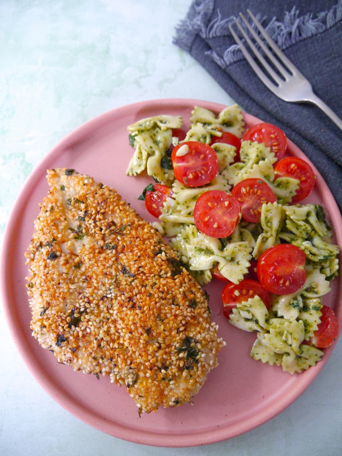 A pink plate topped with sesame crusted chicken breast with a side of pesto pasta salad and sliced cherry tomatoes.