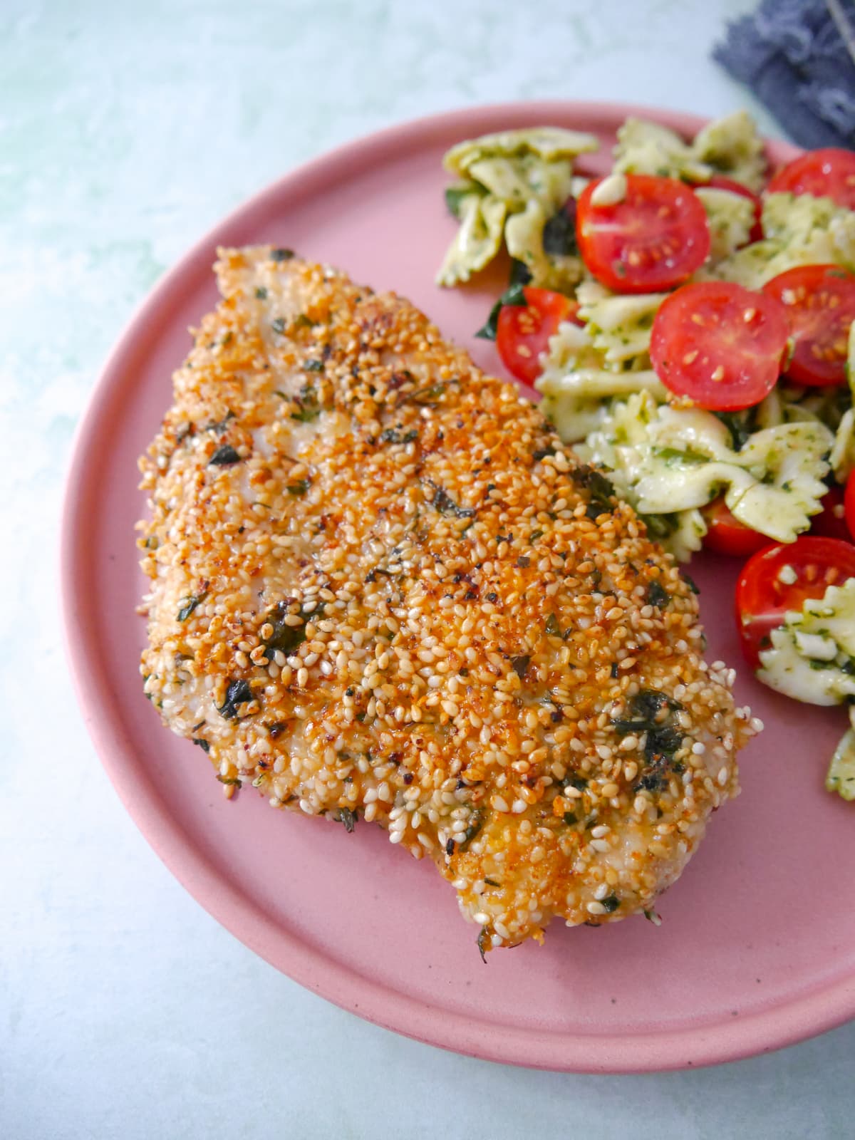 A pink plate topped with sesame crusted chicken breast with a side of pesto pasta salad and sliced cherry tomatoes.