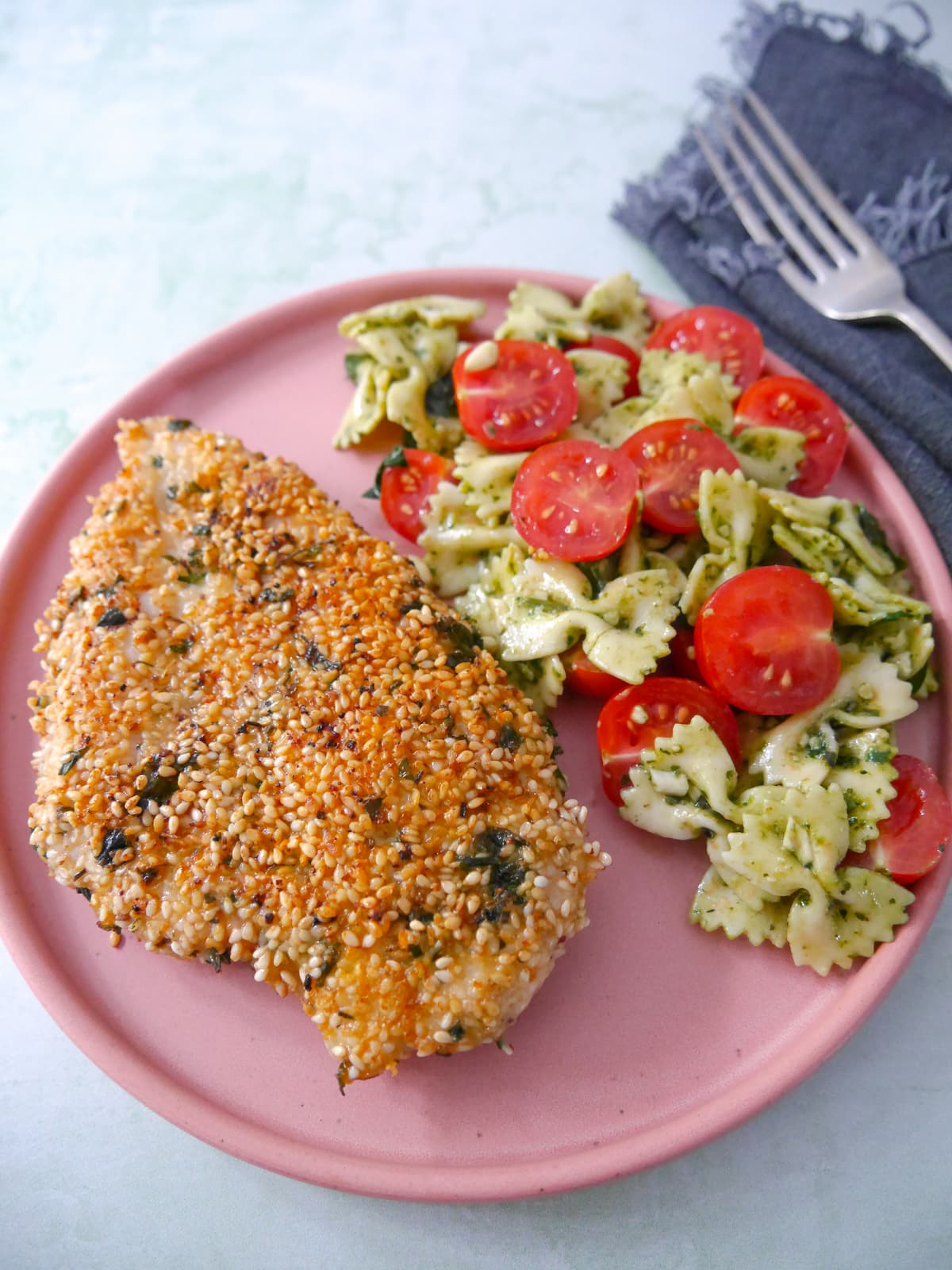 A pink plate topped with sesame crusted chicken breast with a side of pesto pasta salad and sliced cherry tomatoes.