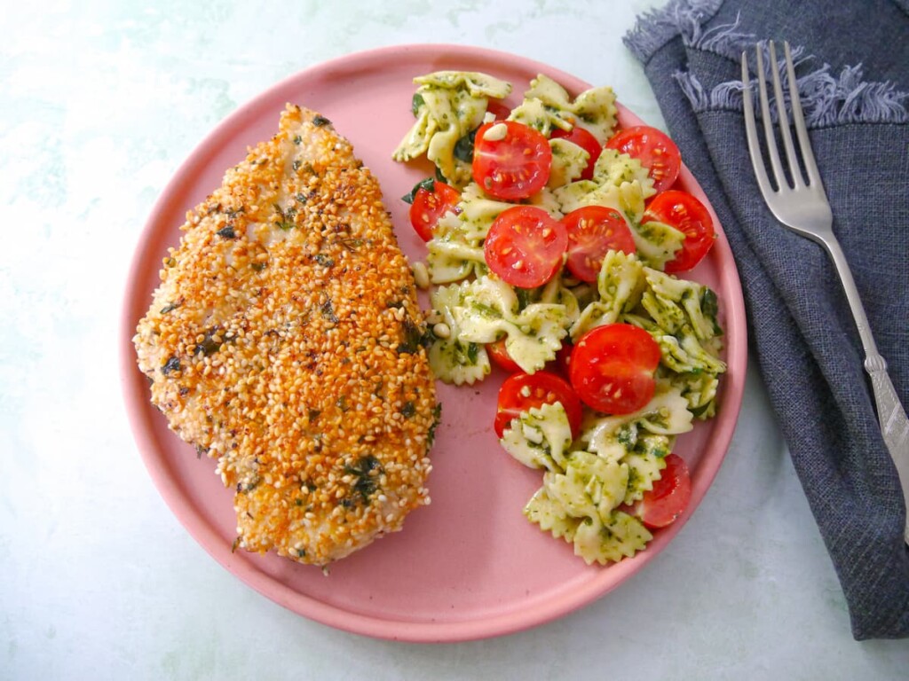 A pink plate topped with sesame crusted chicken breast with a side of pesto pasta salad and sliced cherry tomatoes.