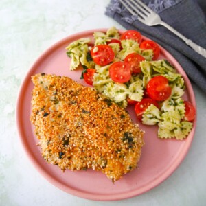 A pink plate with a sesame crusted chicken breast, with a side of tomato and basil pasta salad.