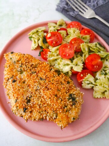 A pink plate with a sesame crusted chicken breast, with a side of tomato and basil pasta salad.