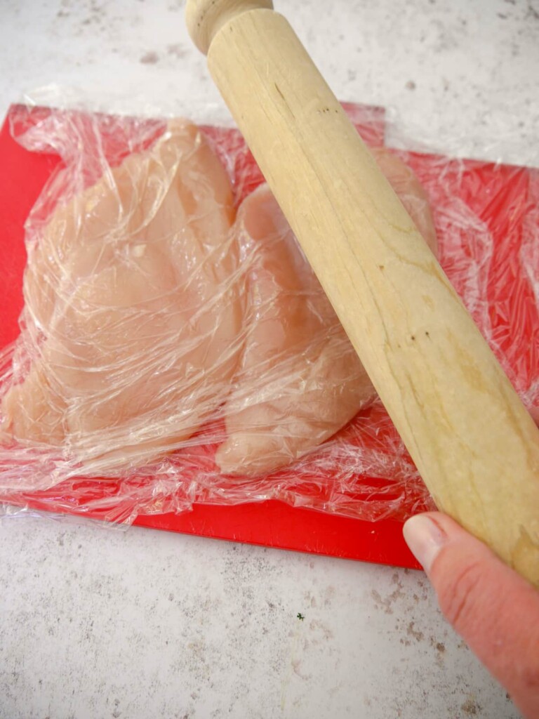 Two chicken breasts covered with clingfilm being gently flattened with a rolling pin.