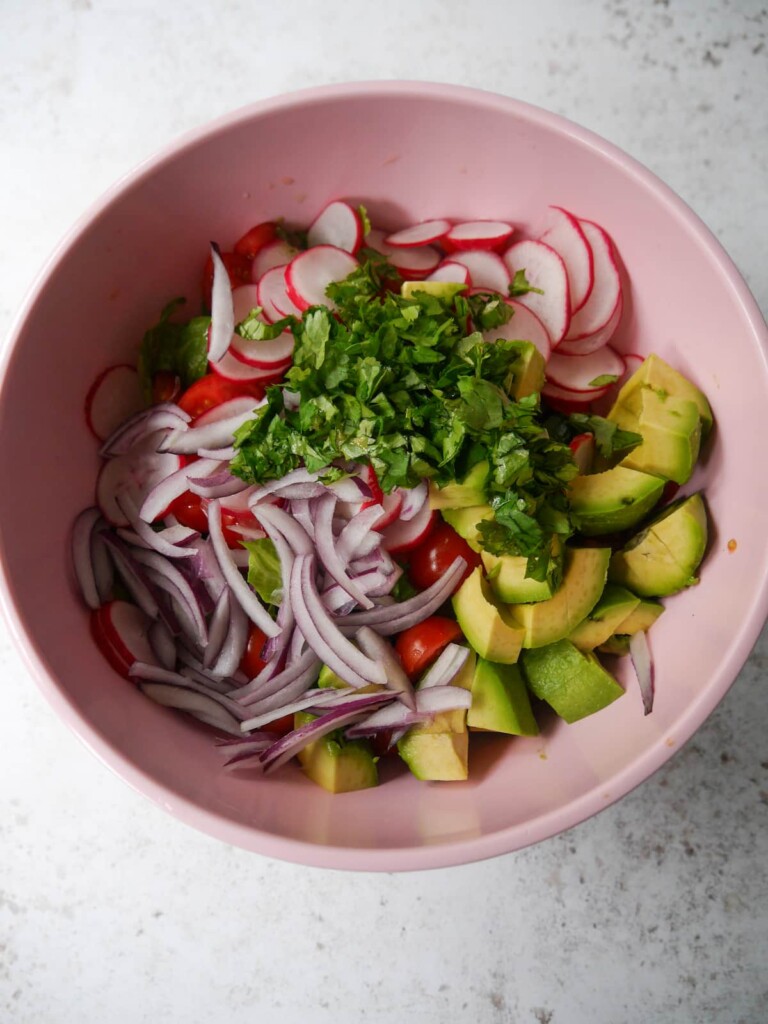 A bowl of salad vegetables including red onion, radish, lettuce leaves, cucumber, tomatoes and avocado, topped with freshly chopped herbs.