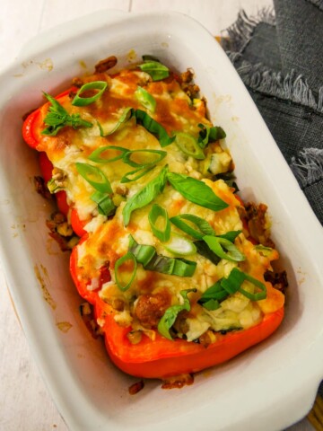 An oven proof dish with 2 halves of stuffed red pepper topped with melted cheese and a garnish of sliced spring onion and shredded basil leaves.