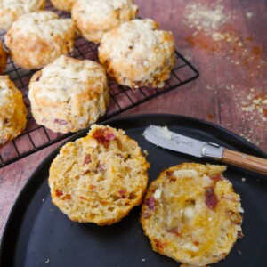 A plate with a cut and buttered cheese bacon tomato scone on top, with a wire rack of scones set alongside.