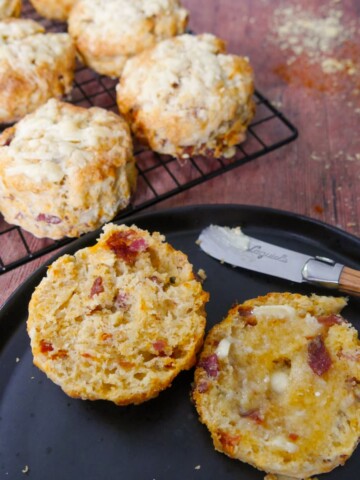A plate with a cut and buttered cheese bacon tomato scone on top, with a wire rack of scones set alongside.