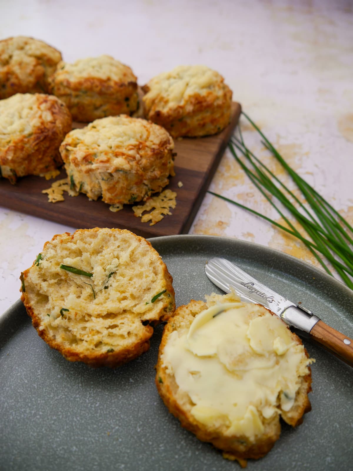 A plate with a halved cheese and chive scone topped with butter. A board of scones is set alongside.