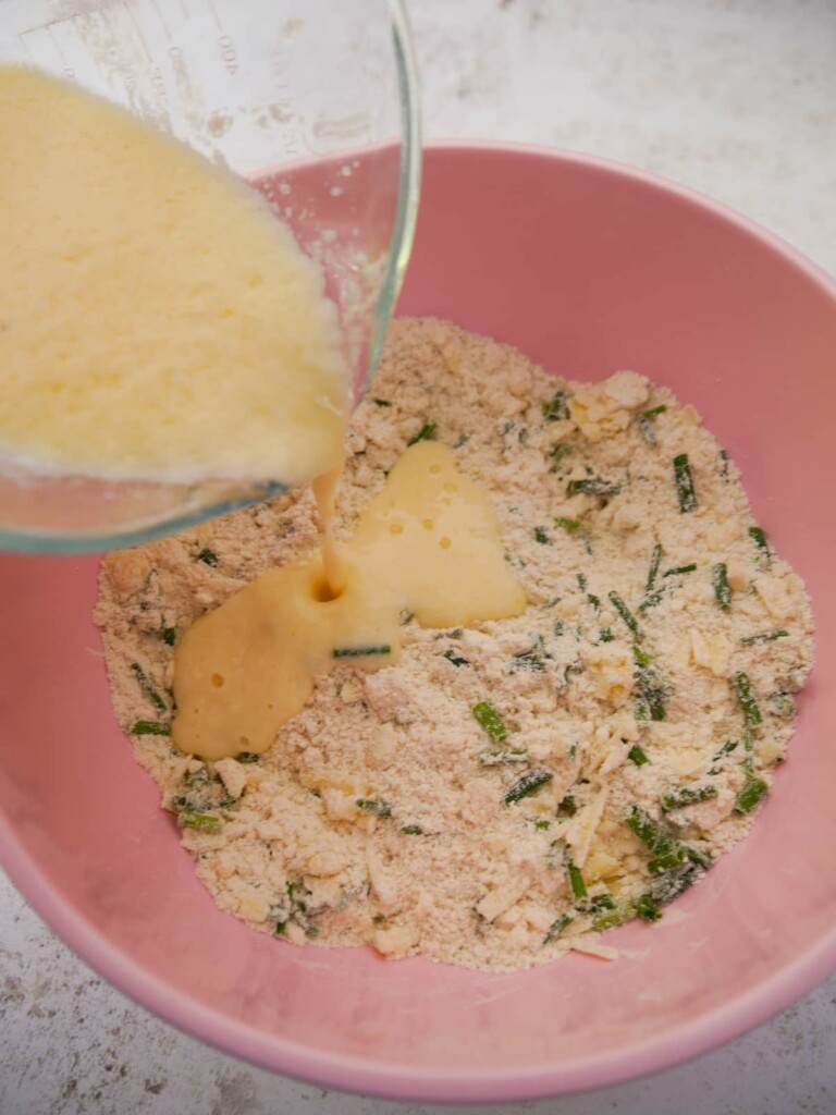 A bowl of dry cheese and chive scone ingredients with beaten egg and buttermilk being added to the bowl.