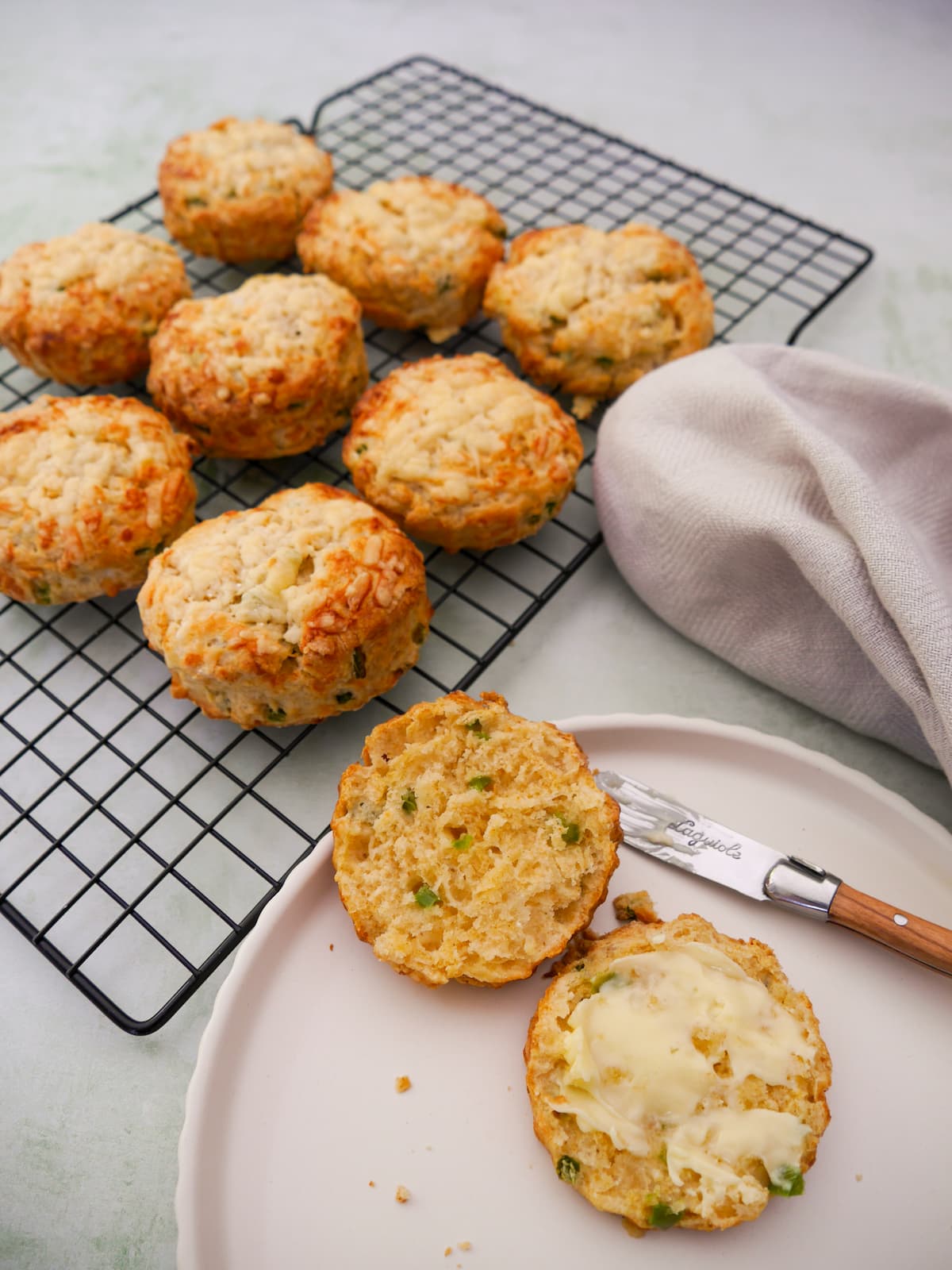 A plate topped with a cheese and jalapeno pepper scone, cut in half and buttered. A wire rack of scones is set alongside.