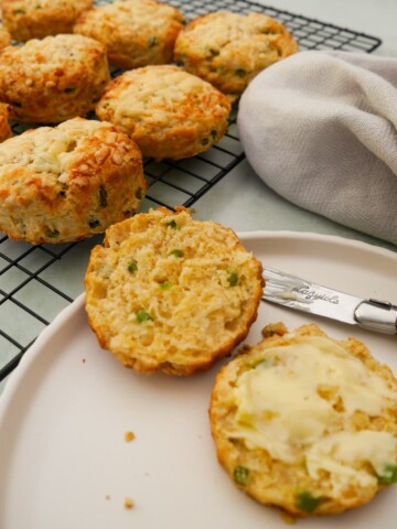 A plate topped with a cheese and jalapeno pepper scone, cut in half and buttered. A wire rack of scones is set alongside.