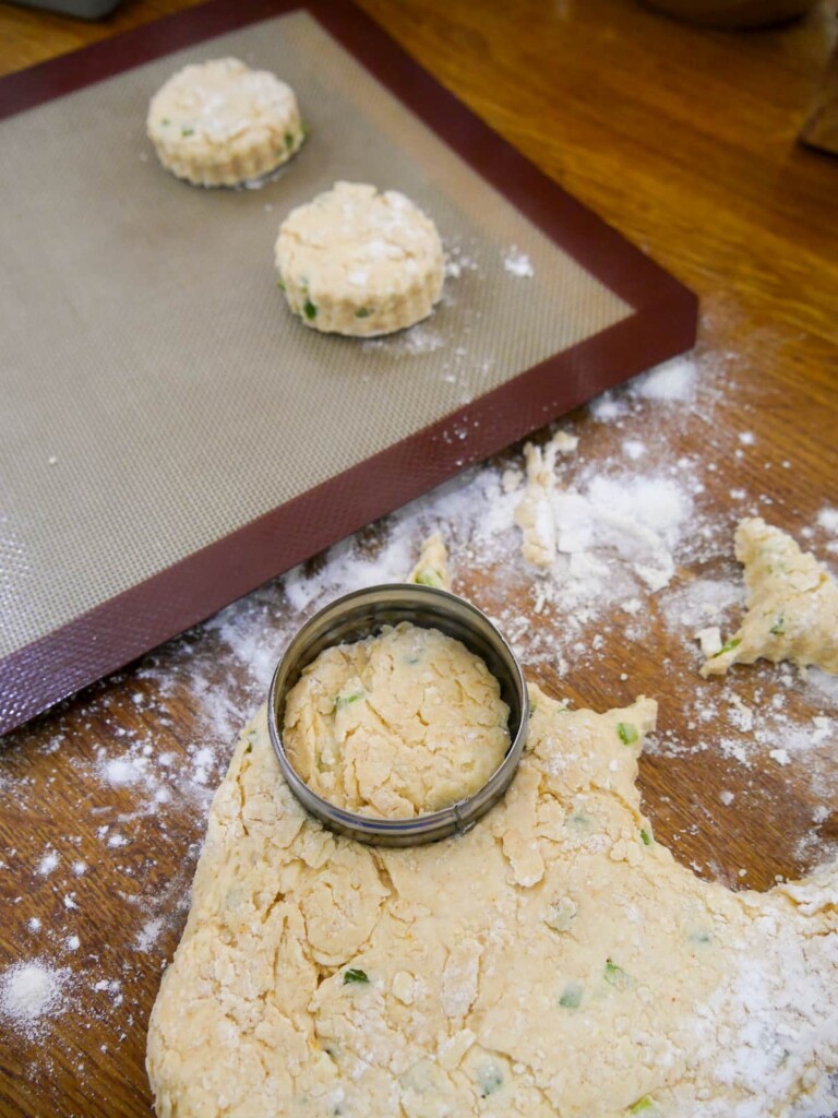 A baking sheet lined with a silicone liner, topped with 2 circular cut scones, with rolled out scone dough and a pastry cutter.
