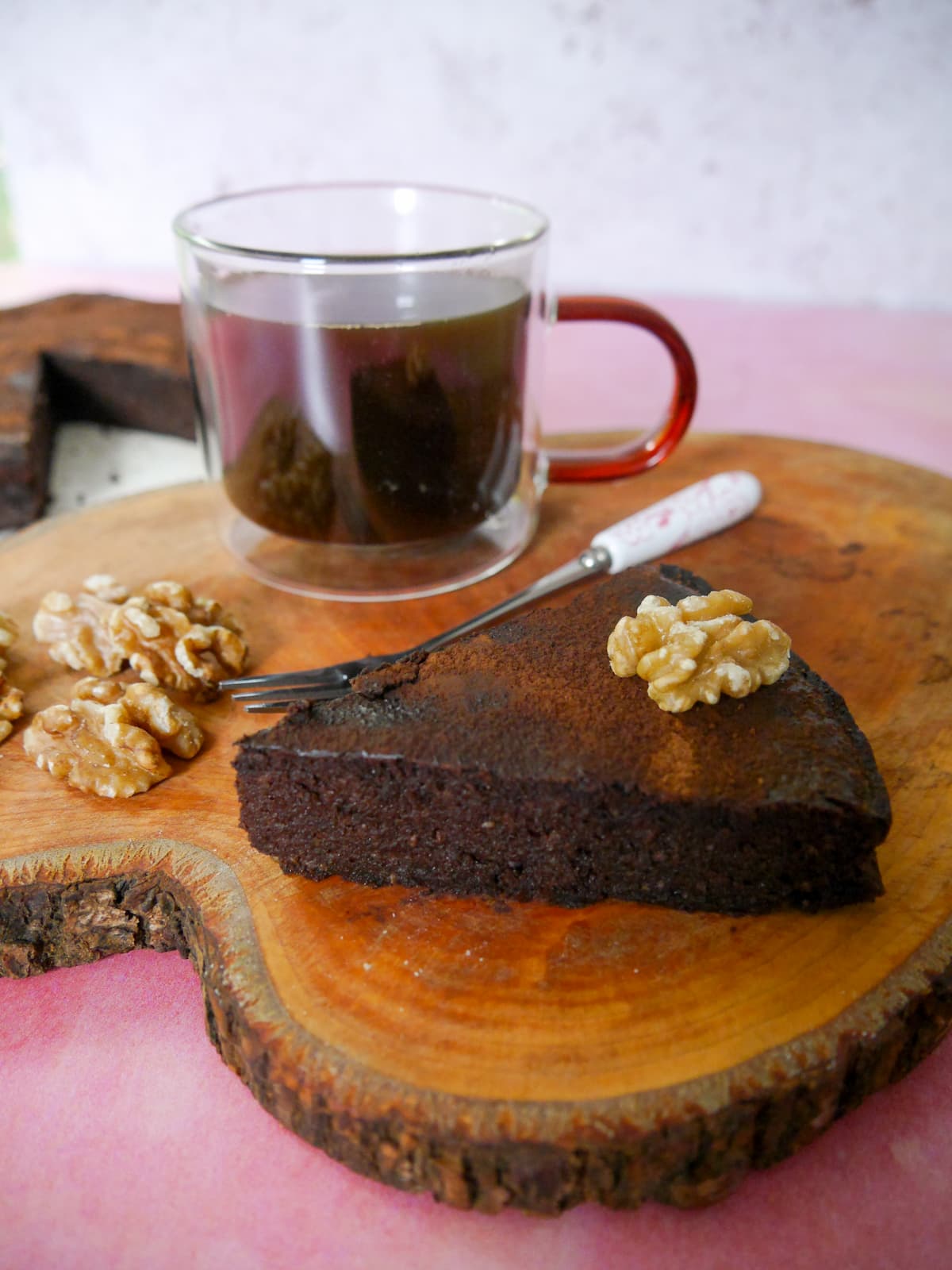 A slice of flourless chocolate coffee walnut cake served with a glass mug of coffee, set on a wooden board, with the whole cake set alongside.