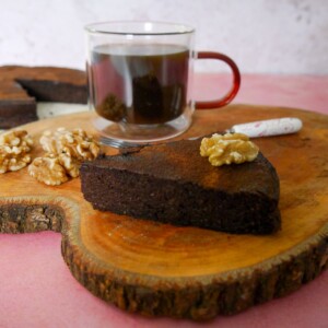 A slice of flourless chocolate coffee walnut cake served with a glass mug of coffee, set on a wooden board, with the whole cake set alongside.