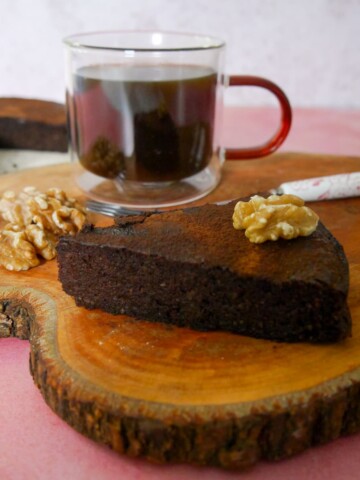 A slice of flourless chocolate coffee walnut cake served with a glass mug of coffee, set on a wooden board, with the whole cake set alongside.