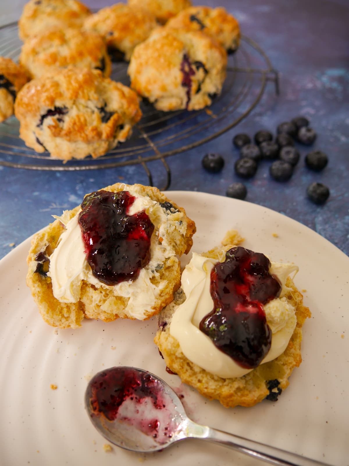A plate of lemon and blueberry scones, topped with clotted cream and blueberry jam. A wire rack of scones is set alongside.